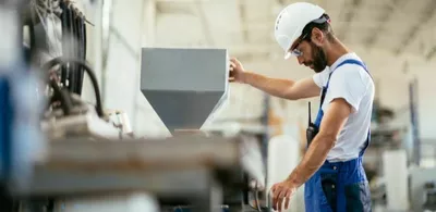 En mann står og arbeider på en fabrikk med hvit hjelm og snekkerbokser. Foto