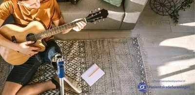 En mann med benprotese sitter på gulvet og spiller gitar. Foto