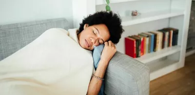 En dame ligger sovende i en sofa med et pledd over seg. Foto