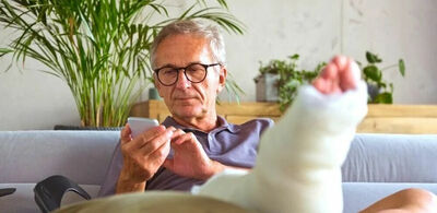 En mann med bandasjert fot ligger i en sofa og ser på telefonen sin. Foto